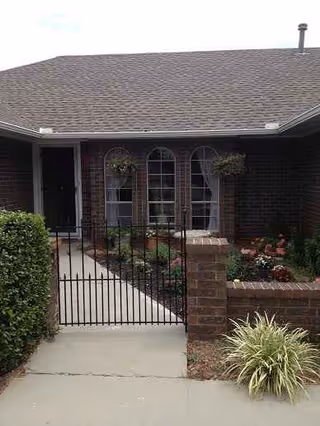 Brick-faced senior living building entrance with arched windows, iron gate, and manicured plants.