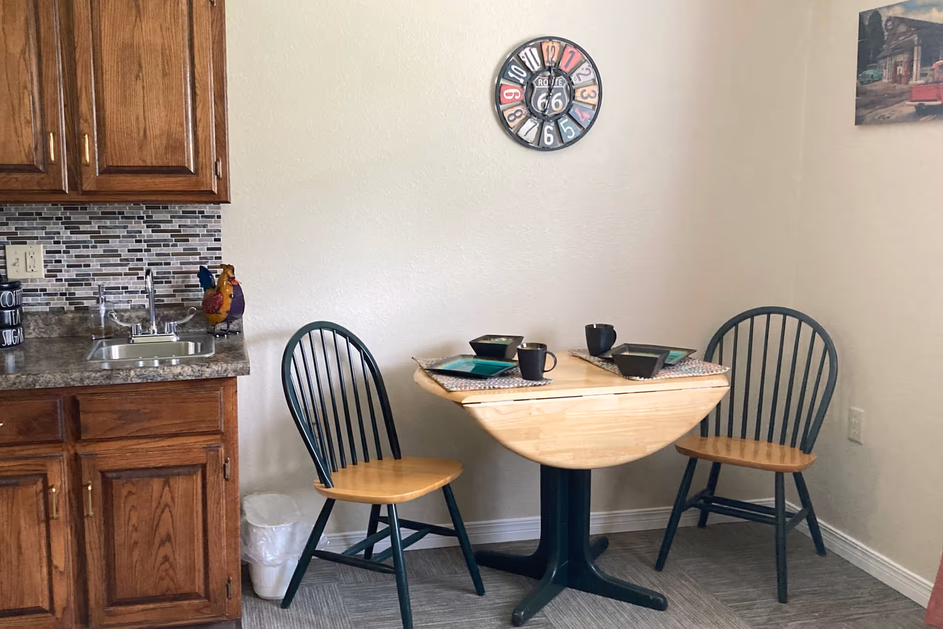 A small dining area with a round wooden table set for two with black mugs and square plates. Two wooden chairs with black backs and seats are positioned on either side of the table. To the left is a kitchen counter with a sink, wooden cabinets, and a decorative rooster figurine. A colorful wall clock hangs on the plain white wall above the table, and a framed picture is partially visible on the right wall.