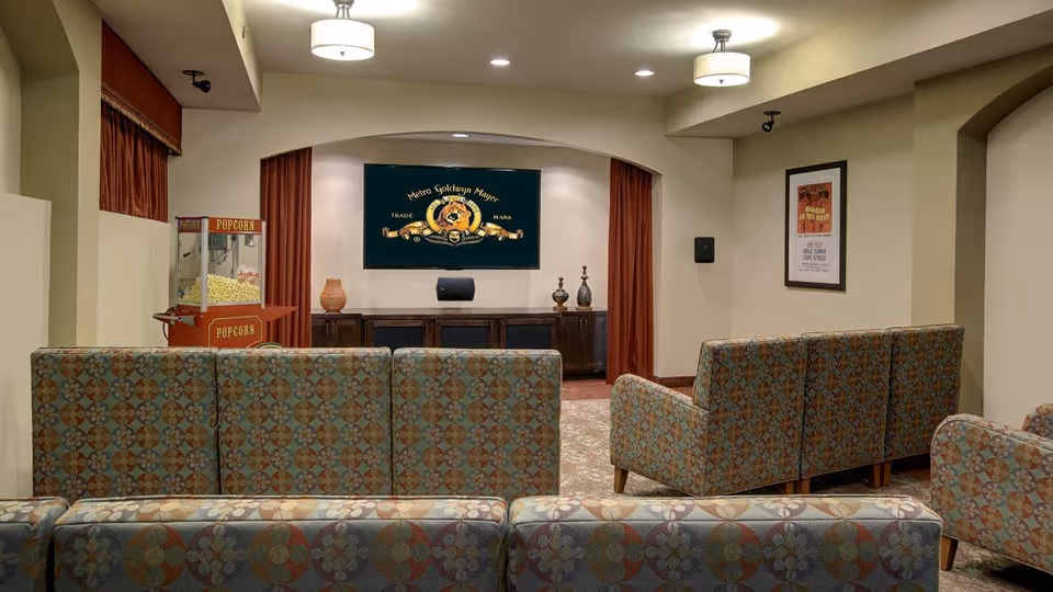 A cozy movie room with patterned upholstered chairs arranged in rows facing a large flat-screen TV mounted on the wall. There is a vintage-style popcorn machine on the left side and decorative vases on a wooden cabinet beneath the TV. The room has warm lighting with ceiling fixtures and framed movie posters on the walls.