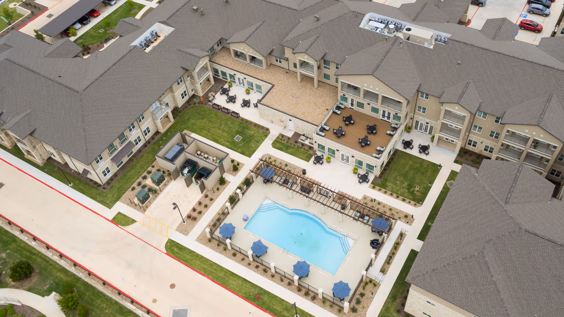 Aerial view of The Enclave at Round Rock Senior Living showing a central outdoor swimming pool surrounded by lounge chairs and umbrellas. The pool area is enclosed by a fence and adjacent to a patio with tables and chairs. Surrounding the pool are multi-story residential buildings with balconies and landscaped green areas. A parking lot is visible in the background.