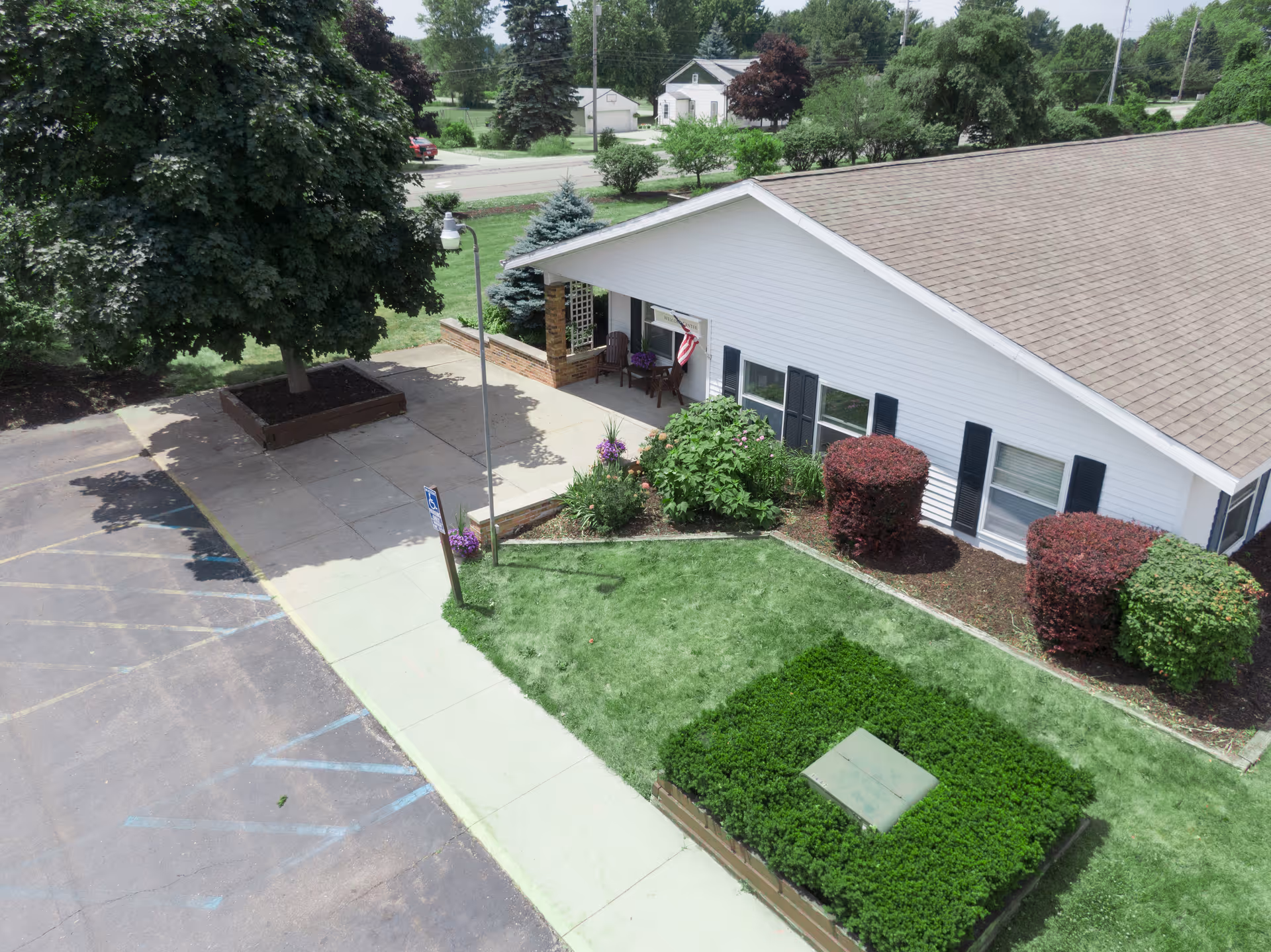 Exterior view of a single-story white building with a brown shingled roof, surrounded by well-maintained landscaping including bushes, a tree, and flower beds. There is a concrete sidewalk and a parking lot with handicap parking spaces visible. A small porch with chairs and an American flag is seen near the entrance.