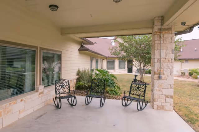Covered outdoor patio area with three black metal rocking chairs, beige stone pillars, and windows looking into the building. The patio overlooks a grassy area with shrubs and trees.