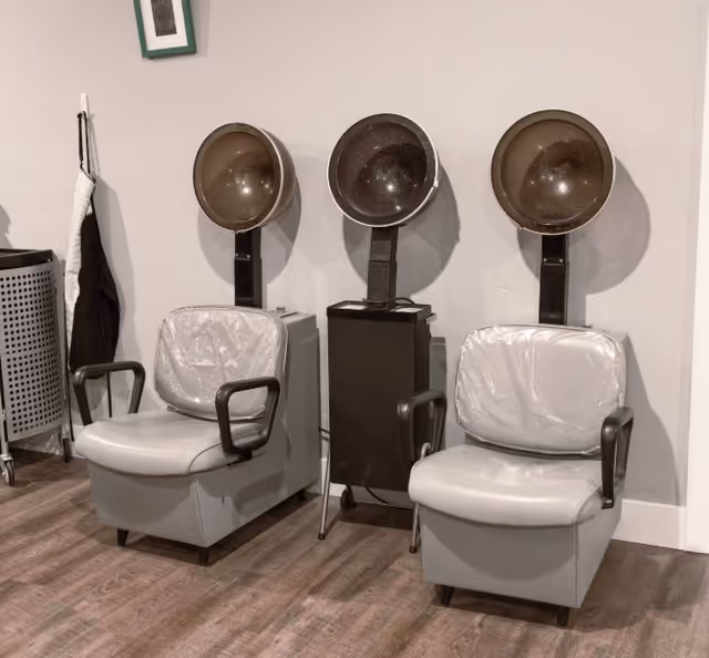 Two vintage salon hair dryers with attached chairs in a room with wooden flooring and a white wall. There is a small framed picture hanging on the wall and a laundry basket with a towel hanging on the left side.