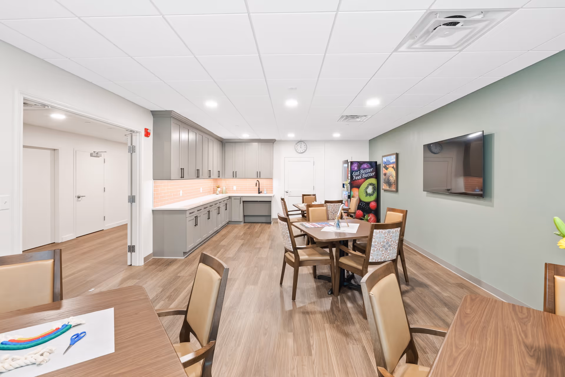 A bright and clean common dining area in a senior living facility with wooden tables and chairs. The room features a kitchenette with gray cabinets, a sink, and a light pink tiled backsplash. A vending machine with colorful fruit images and the text 'Eat Better Feel Better' is visible against a green wall, which also has a mounted flat-screen TV and framed artwork. The floor is wood laminate, and the ceiling has recessed lighting.