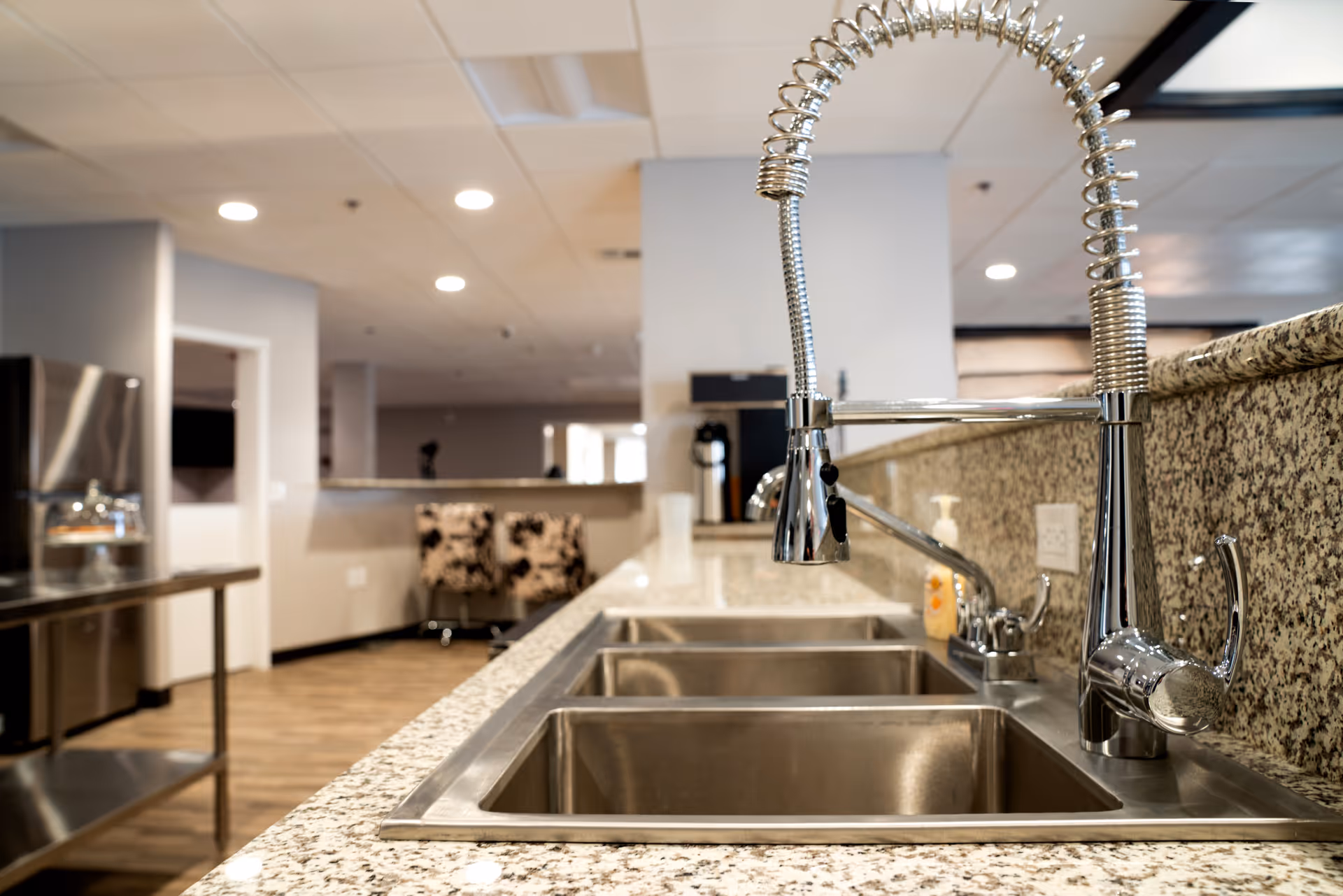 Stainless steel triple sink with a spring-coil faucet set into a granite countertop in a communal kitchen area.