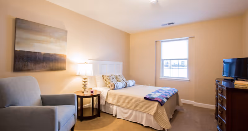 A cozy bedroom with a bed covered in beige bedding and decorative pillows, a colorful quilt at the foot of the bed, a round wooden side table with a lamp, a gray upholstered armchair, a window with blinds, a wooden dresser with a small TV on top, and a large abstract painting on the wall.