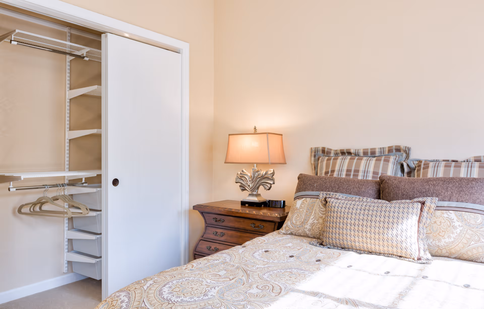 A cozy bedroom featuring a bed with patterned bedding and multiple pillows, a wooden nightstand with a decorative lamp, and an open closet with shelves and hanging space.