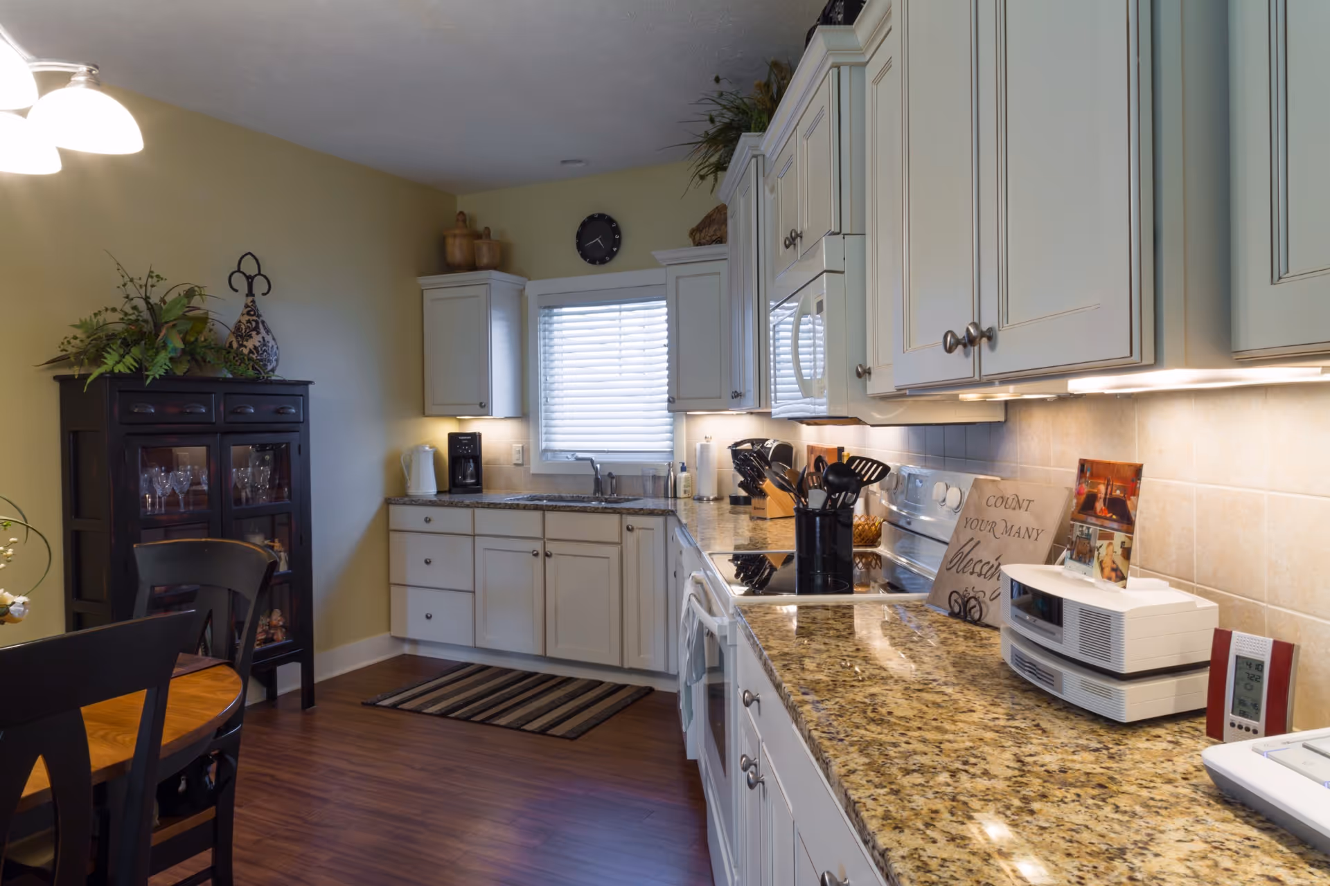 A well-lit kitchen with white cabinets, granite countertops, and various kitchen utensils. There is a window with blinds above the sink, a coffee maker on the counter, and a dining table with chairs to the left. The floor is wooden, and there is a decorative cabinet with glassware and plants on top.