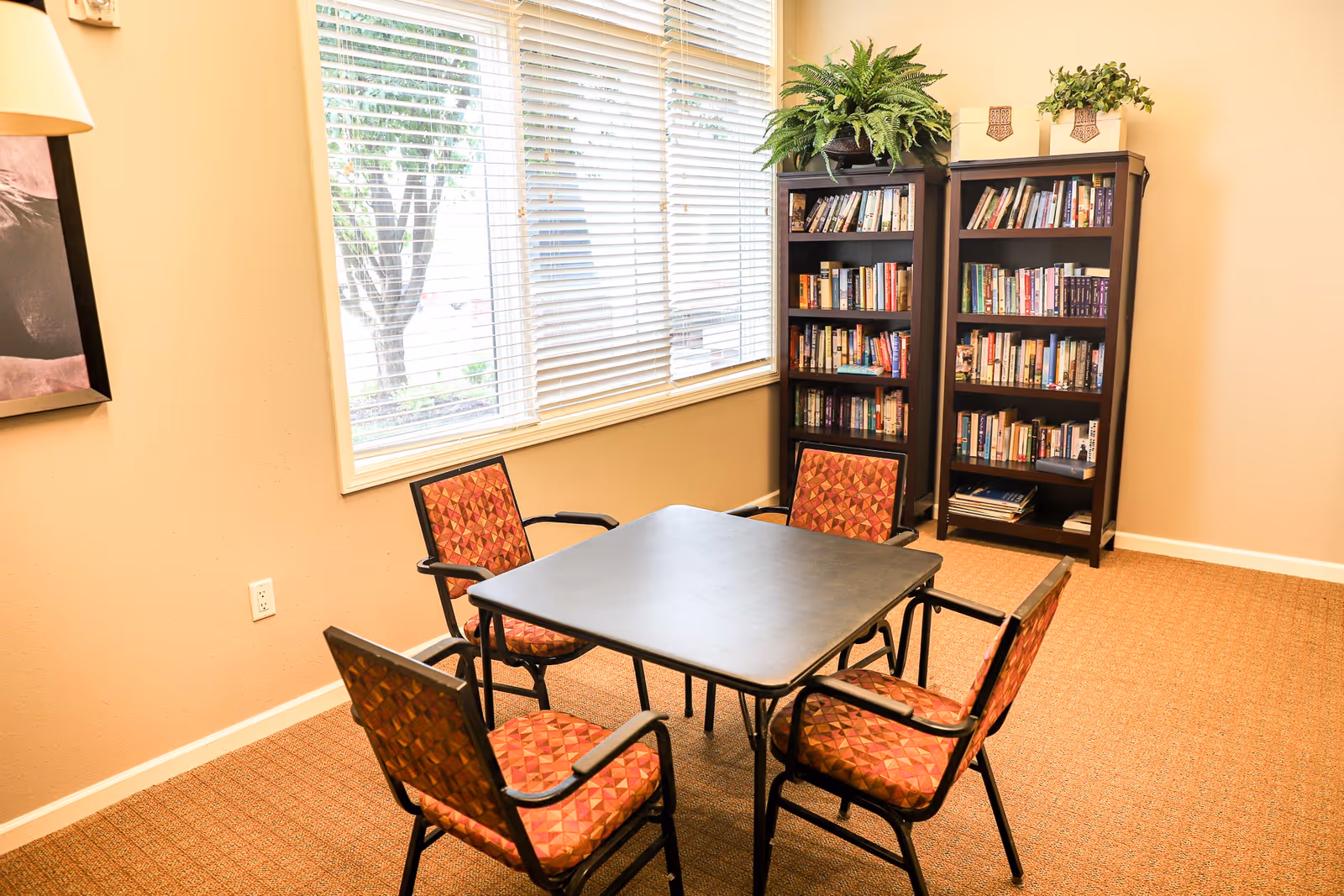 Small common room with a square table surrounded by four patterned chairs, bookshelves and a large window.