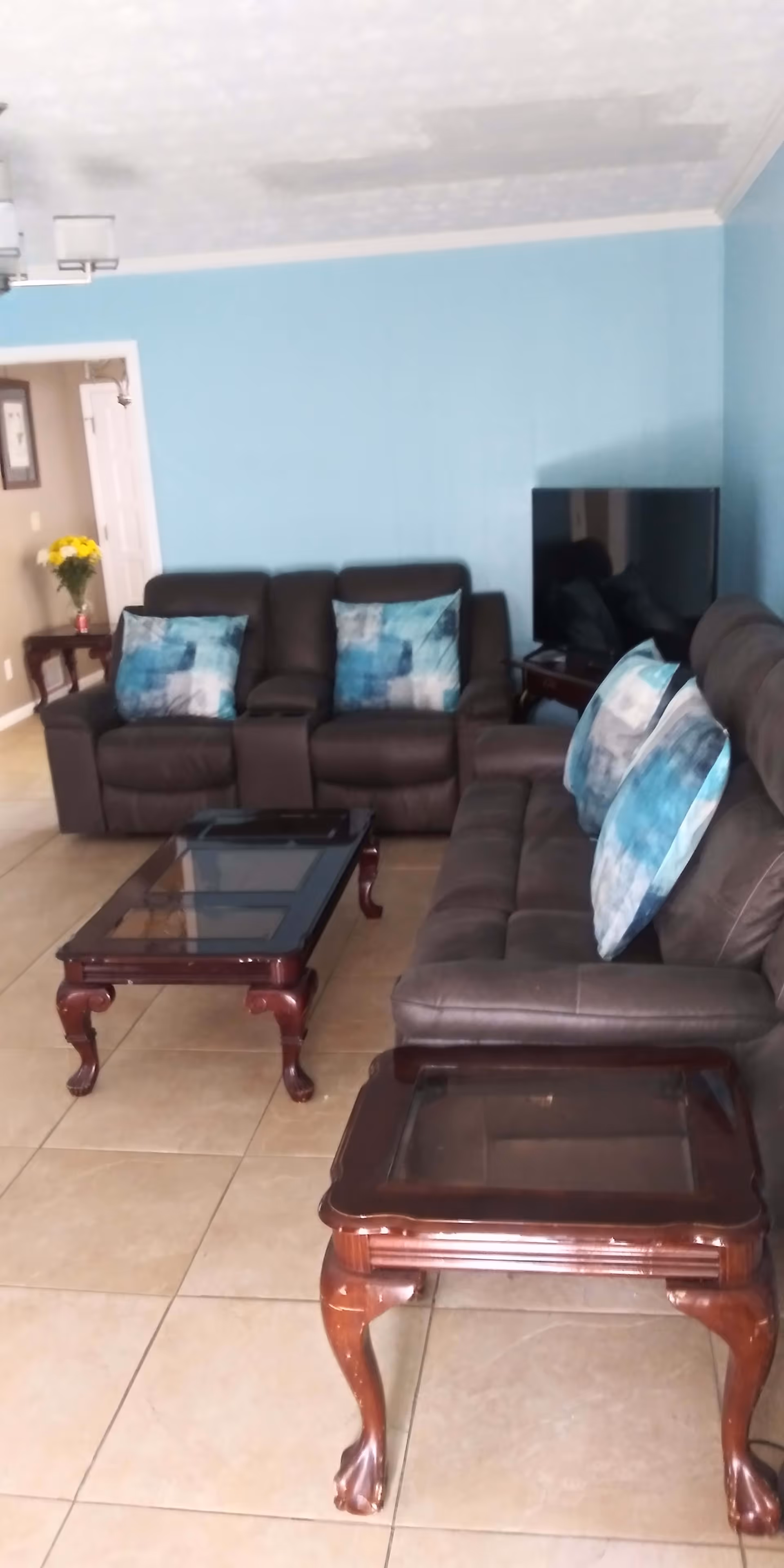 A living room with two dark brown leather sofas, each adorned with blue and white patterned throw pillows. There are two wooden glass-top tables with carved legs, one coffee table in front of the sofas and one side table. A flat-screen TV is placed on a small wooden stand against a light blue wall. The floor is tiled, and there is a doorway leading to another room with a vase of yellow flowers visible on a small table.