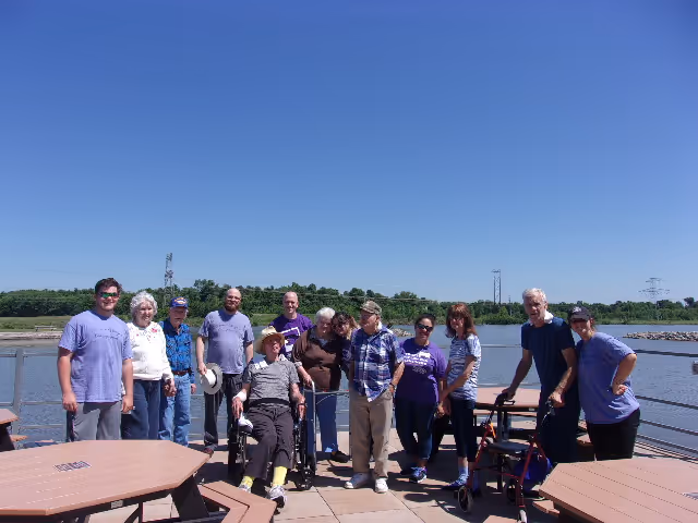 A group of elderly people and caregivers standing together on a wooden deck by the water on a sunny day. Some individuals are using wheelchairs and walkers. Picnic tables are visible in the foreground, and there is a clear blue sky with greenery in the background.
