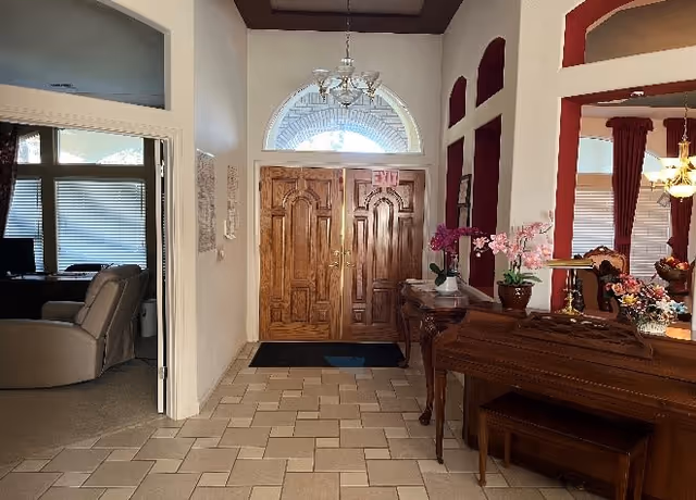 Interior view of a senior living facility entrance area with double wooden doors featuring an exit sign above. The floor is tiled with a beige and light brown pattern. To the right, there is a wooden piano with a bench and decorative flowers on top. The walls have red accent niches with arches, and a chandelier is visible hanging from the ceiling. To the left, there is an open doorway leading to a room with a recliner chair and a window with blinds.