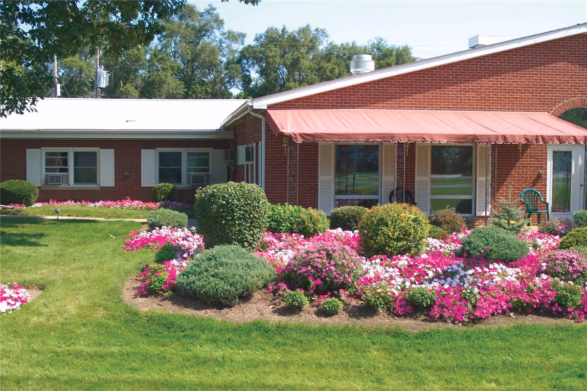 Front of a single-story red-brick building with a red awning, patio chairs, and vibrant flower beds and lawn.