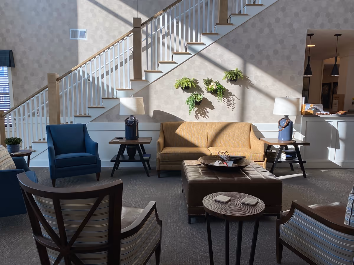 Sunlit communal seating area with a sofa, armchairs, ottoman, side tables, and a staircase in the background.