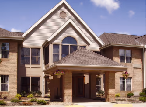 Exterior front view of a two-story senior living facility building with a covered entrance, brick and beige siding, multiple windows, and hanging flower baskets under a clear blue sky.