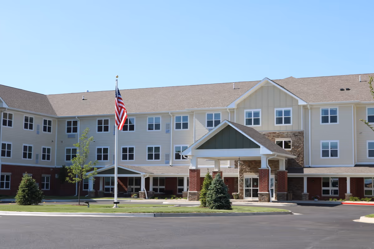 Three-story senior living facility building with a covered entrance, American flag on a flagpole, and landscaped front.