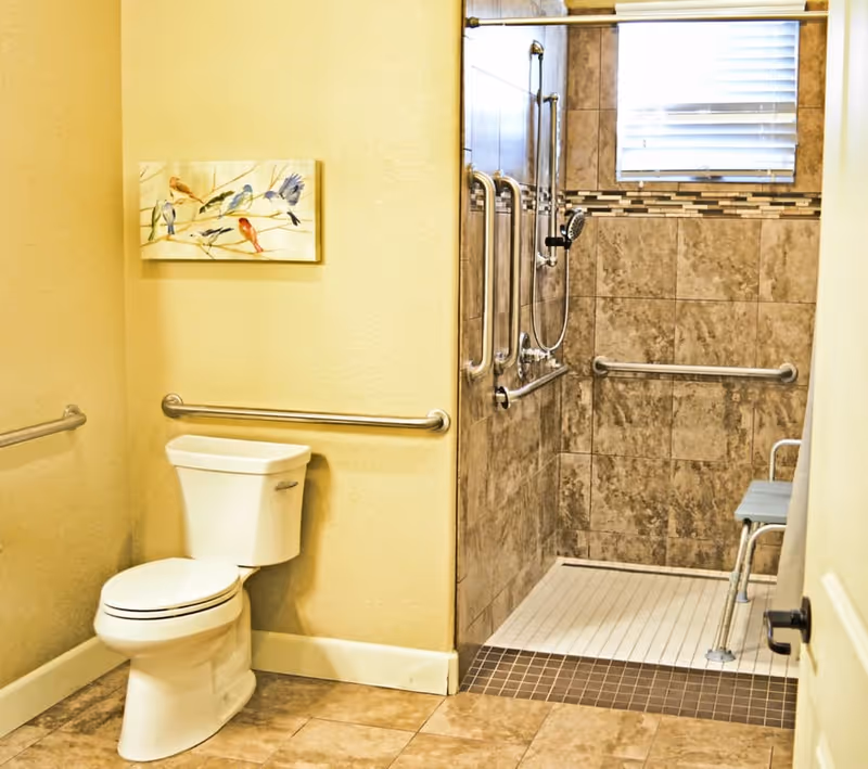 A bathroom with beige walls and tiled floor featuring a white toilet with metal grab bars on the walls. To the right, there is a walk-in shower with brown and beige tiles, multiple grab bars, a handheld showerhead, a small window with blinds, and a shower chair.