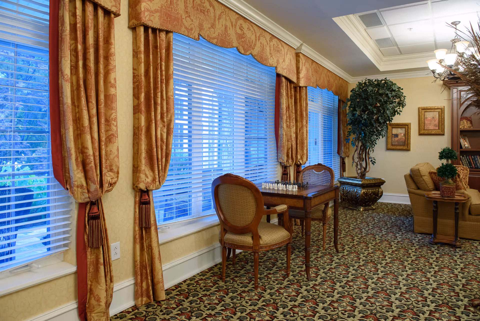 A cozy interior room with large windows covered by patterned gold and red curtains. A wooden table with a chess set and two upholstered chairs are positioned near the windows. The room features a patterned carpet, a large potted plant, framed artwork on the wall, and a comfortable seating area with a sofa and side table.