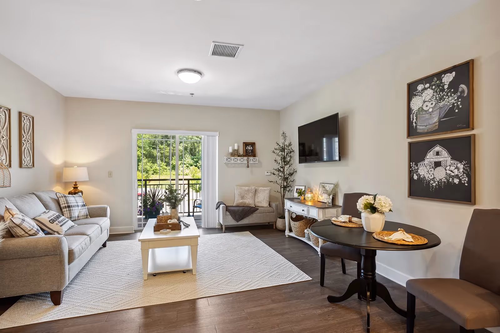 A bright and cozy living room with a gray sofa adorned with plaid and decorative pillows, a white coffee table on a light-colored rug, a small loveseat near a sliding glass door leading to a balcony with greenery outside, a wall-mounted flat-screen TV, a console table with framed pictures and candles, a round dining table set for two with a flower centerpiece, and two framed floral artworks on the wall.