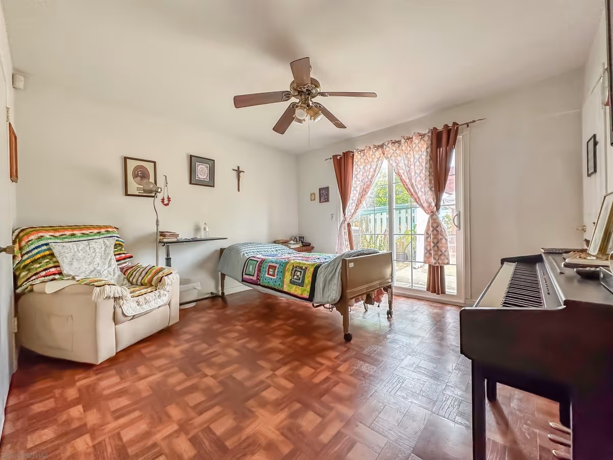 A bright bedroom with a single bed covered with a colorful quilt, a beige armchair with a multicolored crocheted blanket, a ceiling fan, framed pictures and a crucifix on the wall, a sliding glass door with patterned curtains leading to an outdoor area, and a piano on the right side.