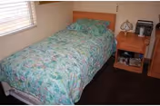 Small bedroom with a single bed covered in a green floral comforter beside a wooden nightstand and a window.