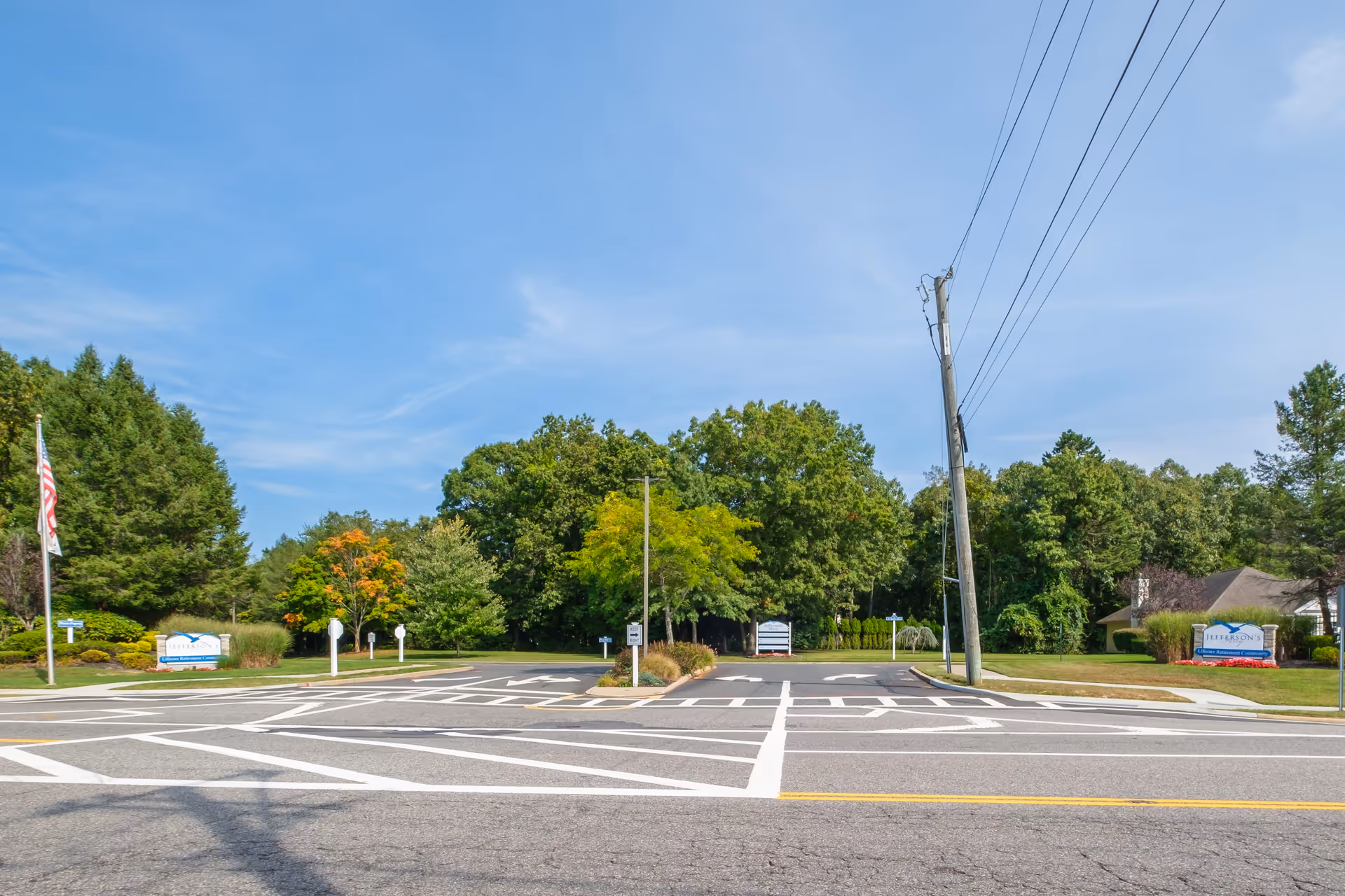 Entrance to a senior living community with landscaped grounds, signage, an American flag, and a road crossing in front.