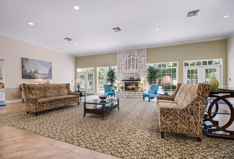 Spacious lounge with patterned sofas, coffee table, stone fireplace, and large windows overlooking greenery.