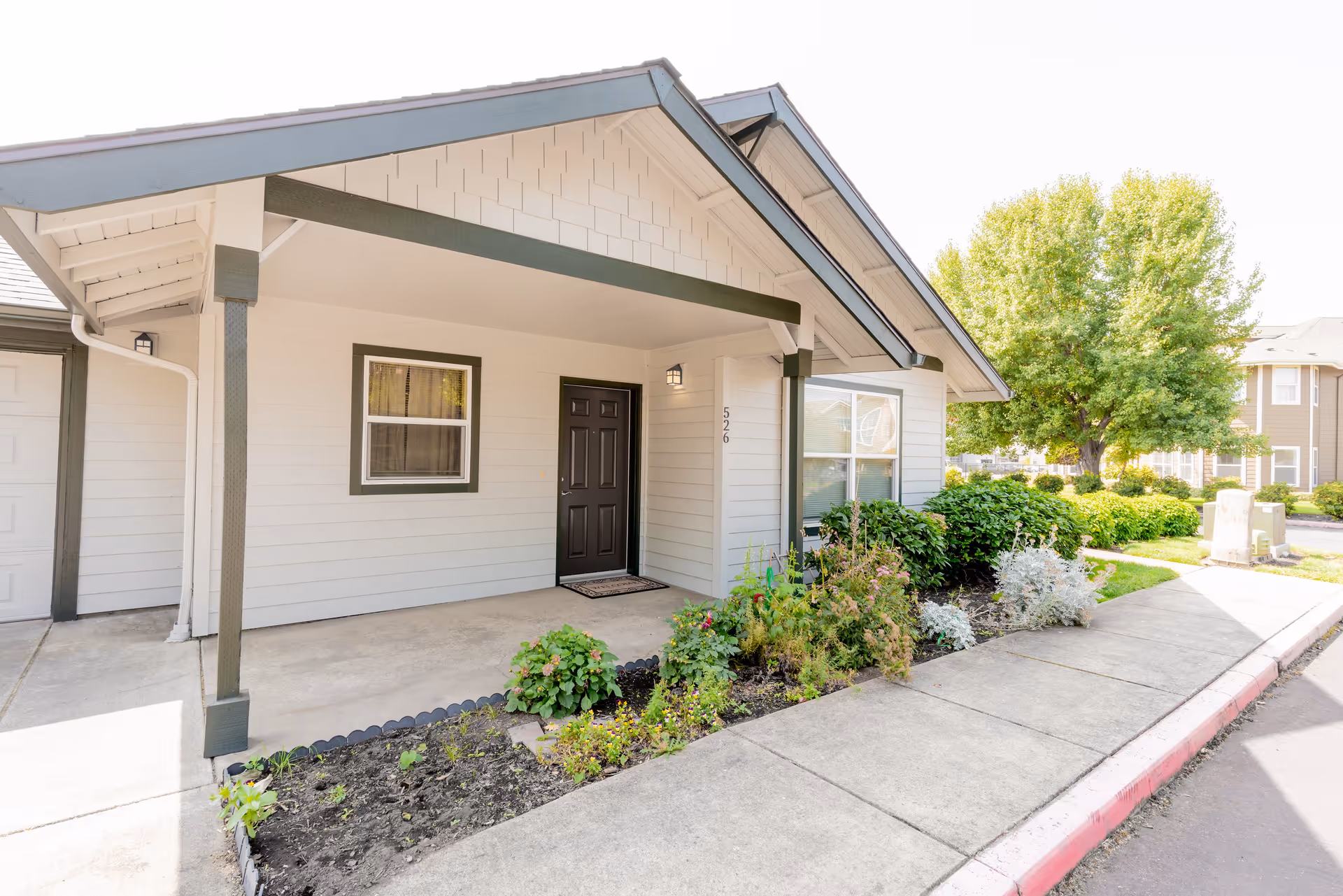 Exterior view of a senior living facility unit with a covered porch, a black front door, a window, and a small garden bed with plants along the sidewalk. The building is light-colored with dark trim, and there are trees and other buildings visible in the background.