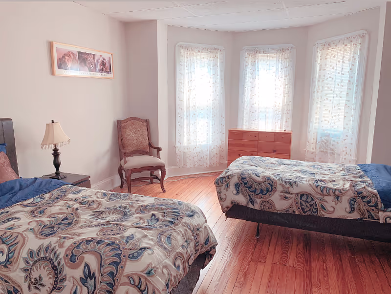 Sunlit bedroom with two twin beds, a wooden dresser, an upholstered chair, and three tall windows with sheer curtains.