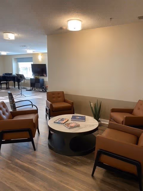 A cozy common area with four brown leather armchairs arranged around a round white marble coffee table with a black base. There are magazines on the table and a small green plant next to it. In the background, there is a piano, a TV on a stand, and large windows letting in natural light. The room has wood flooring and beige walls with ceiling lights.