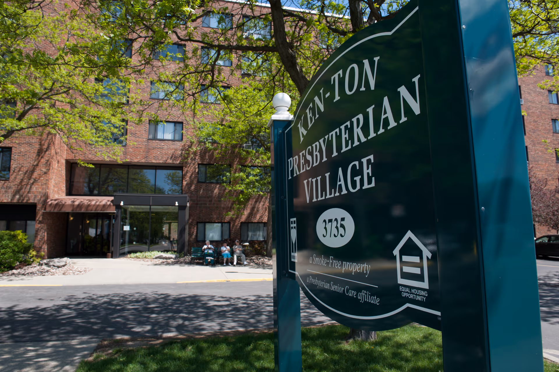 A large green sign reading 'Ken-Ton Presbyterian Village' stands in front of a brick senior living building with a driveway and people seated near the entrance.
