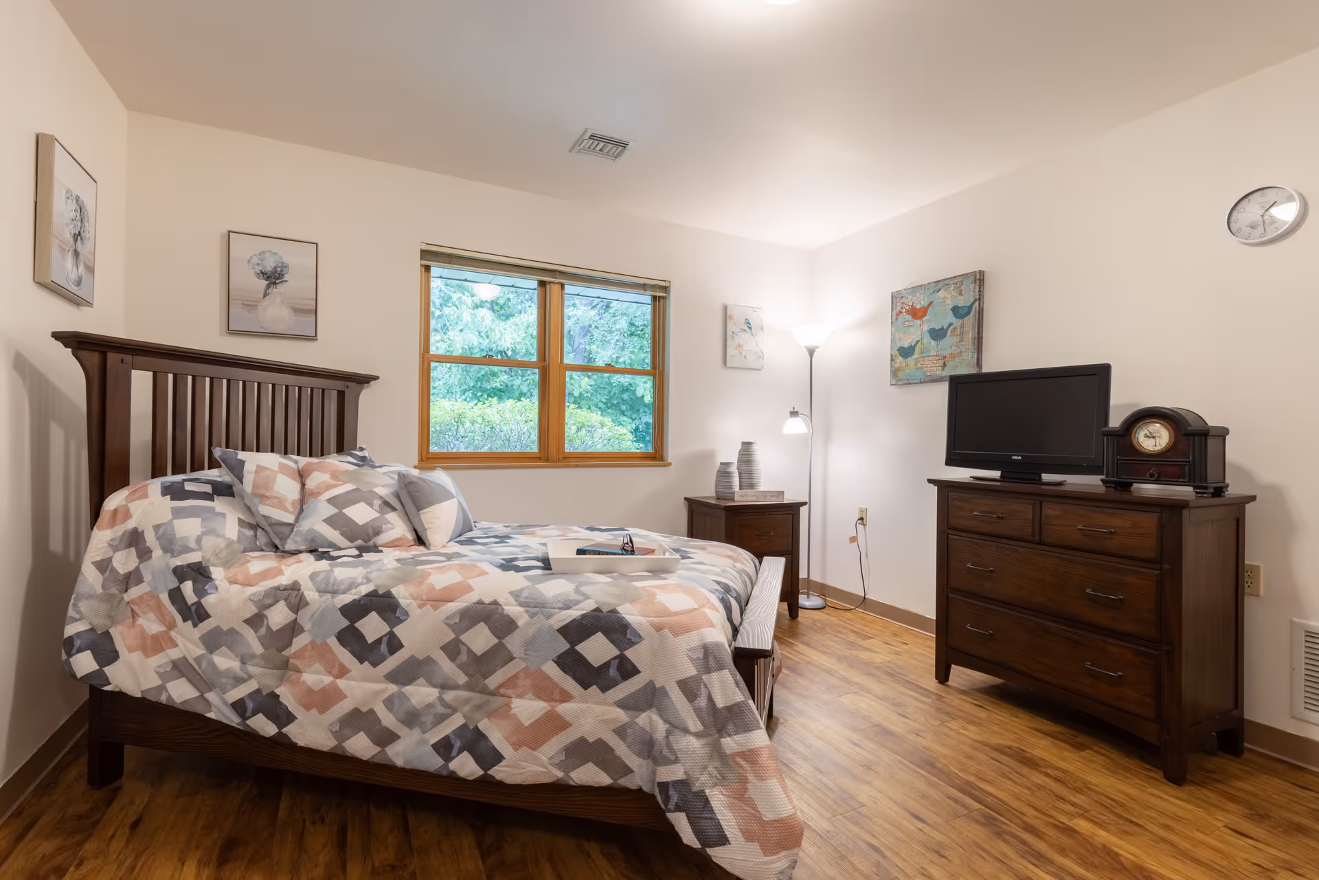 A cozy bedroom with a wooden bed frame and a patterned bedspread in shades of gray, white, and peach. The room has wooden flooring, a window with a view of greenery outside, two framed pictures on the walls, a wooden dresser with a TV and a clock on top, a nightstand with decorative vases, and a floor lamp providing warm lighting.