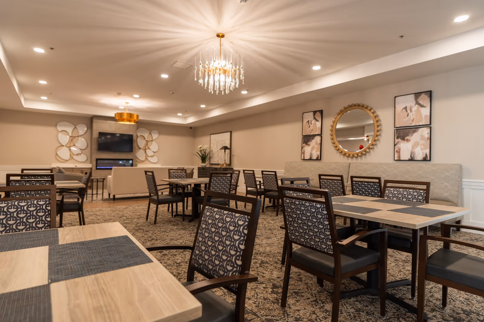 A spacious and well-lit dining area in a senior living facility with multiple wooden tables and patterned chairs. The room features a carpeted floor, modern chandeliers, decorative wall art, a round mirror, and a seating area with sofas and a wall-mounted TV above a fireplace.