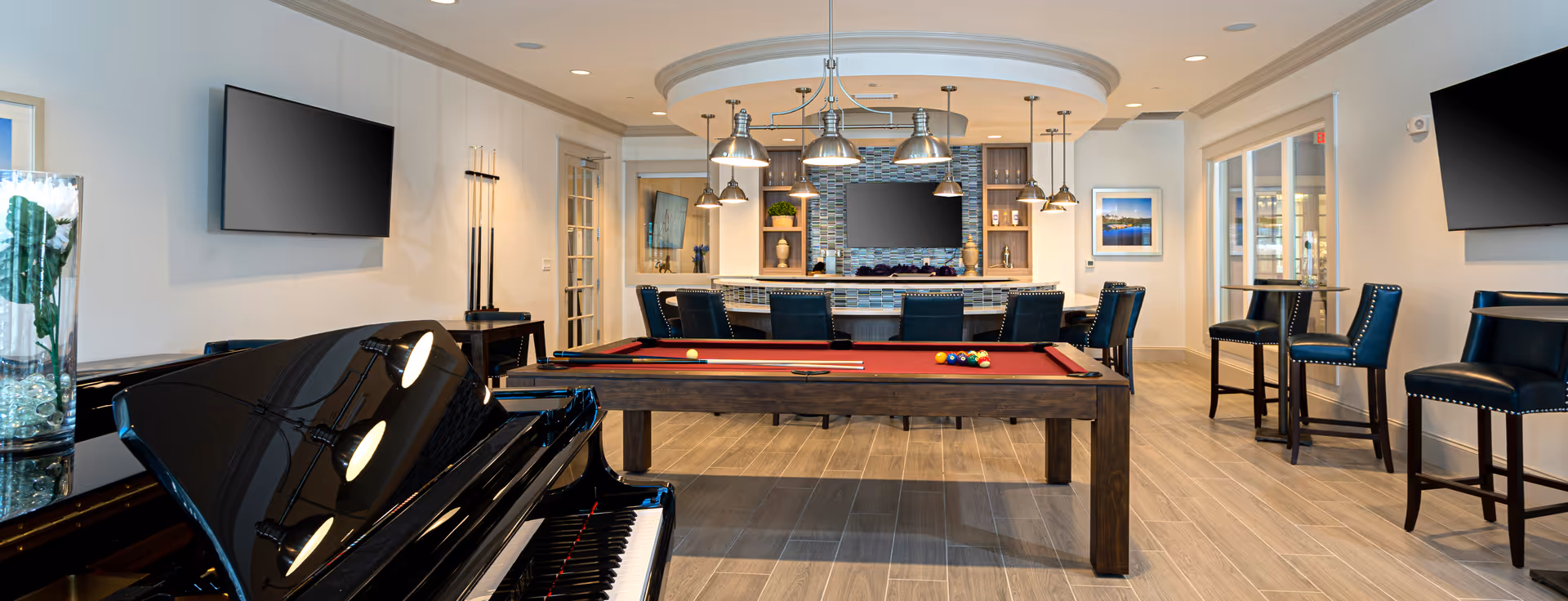 A spacious and well-lit recreational room featuring a red felt pool table with pool cues and balls, a black grand piano, multiple flat-screen TVs mounted on the walls, high bar chairs around a curved bar counter with pendant lights, and additional seating with small tables and chairs. The room has wood-look tile flooring and light-colored walls with crown molding.