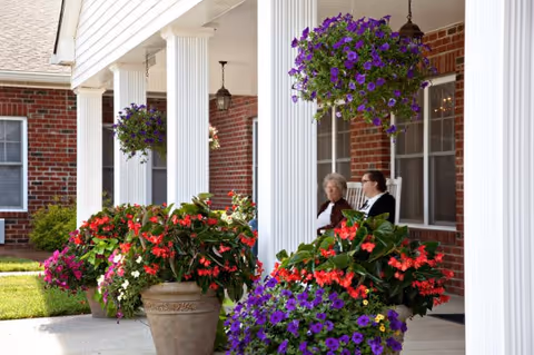 Two elderly women sitting and conversing on a porch of a brick building with white columns, surrounded by large pots of colorful flowers and hanging flower baskets.
