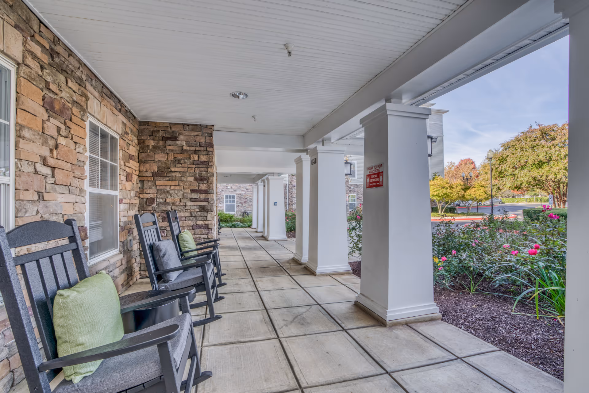 Covered front porch with rocking chairs along a stone wall and white columns overlooking landscaped gardens.