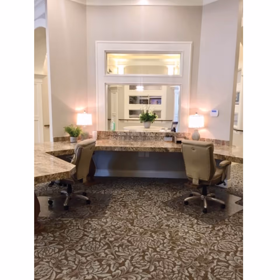 Interior reception area with a marble countertop desk, two rolling chairs, table lamps, potted plants, and patterned carpet.