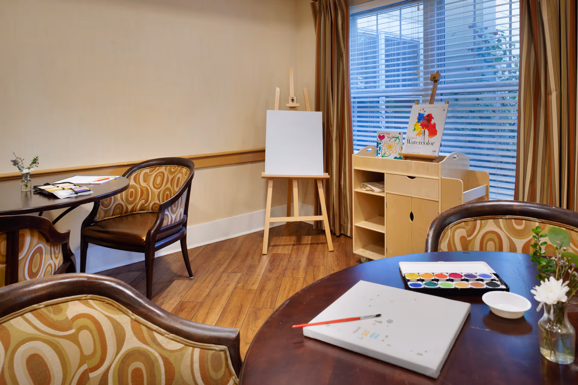 A bright activity room with round tables and patterned chairs, an easel, art supplies, and a large window with blinds.