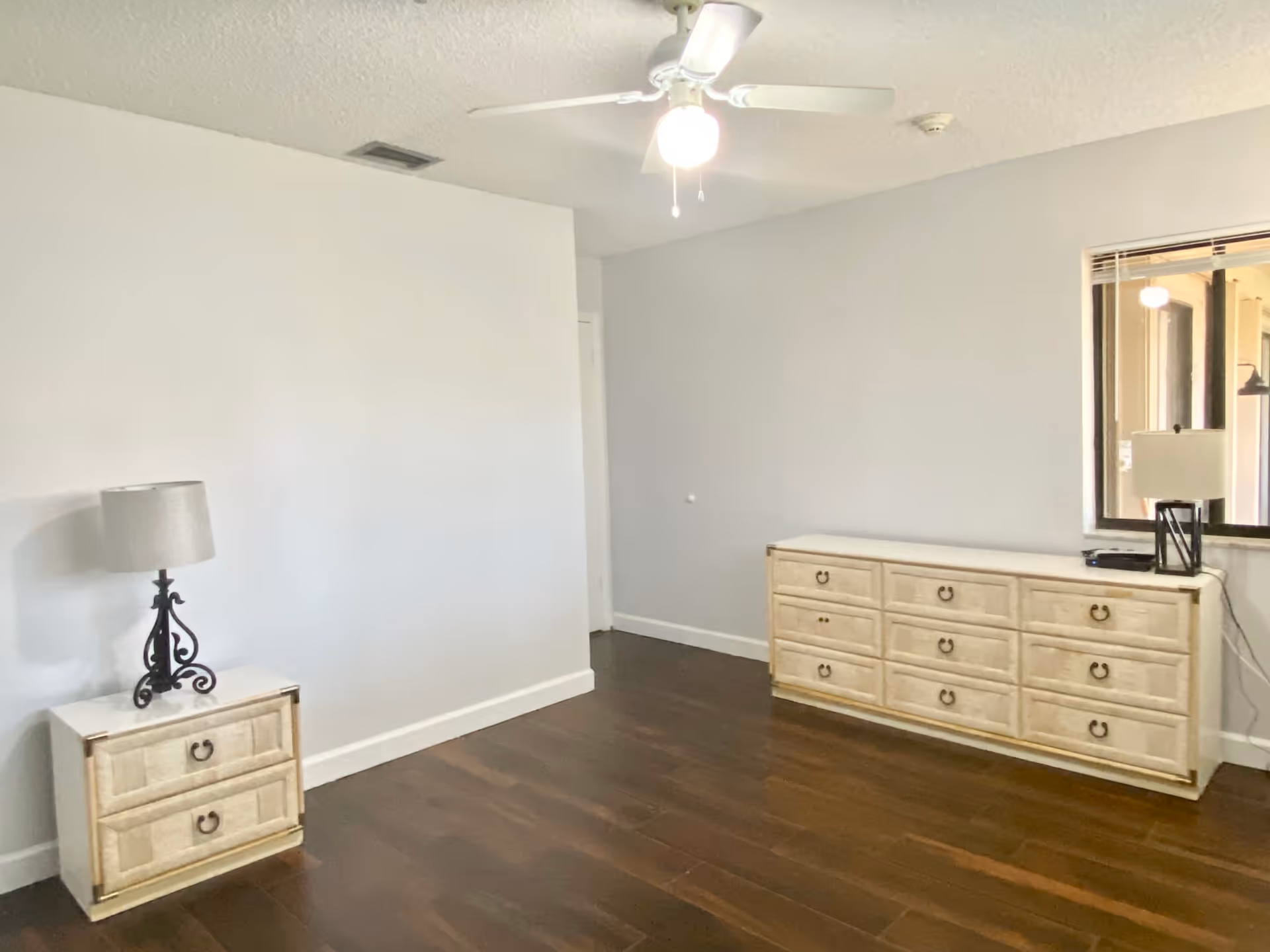 A simple room with light gray walls and dark wooden flooring. There is a small nightstand with a decorative lamp on the left side and a long dresser with multiple drawers on the right side. A ceiling fan with a light is mounted on the ceiling, and a window on the right wall shows an adjacent room with another lamp.