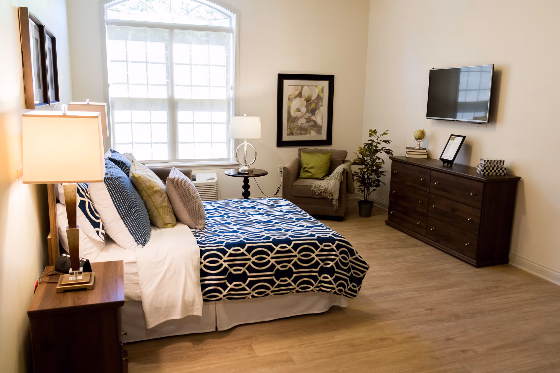 A well-lit bedroom with a large window covered by blinds. The room features a bed with a blue and white patterned blanket and multiple pillows, a wooden nightstand with a lamp, a small round table with a lamp, an armchair with a green cushion and a throw blanket, a framed floral artwork on the wall, a potted plant, a wooden dresser with books, a globe, and a framed picture, and a flat-screen TV mounted on the wall.