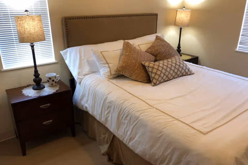 A neatly made bed with white and beige bedding and multiple decorative pillows in a bedroom. There are two bedside tables, each with a lamp and one with a white mug with brown spots. Two windows with blinds are visible on either side of the bed.