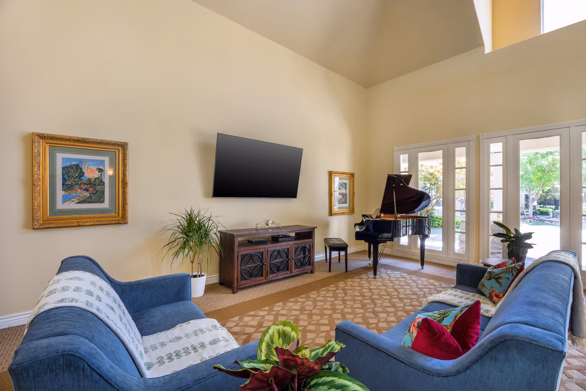 Bright living room with blue sofas facing a wall-mounted TV, a grand piano by tall windows, and framed artwork.