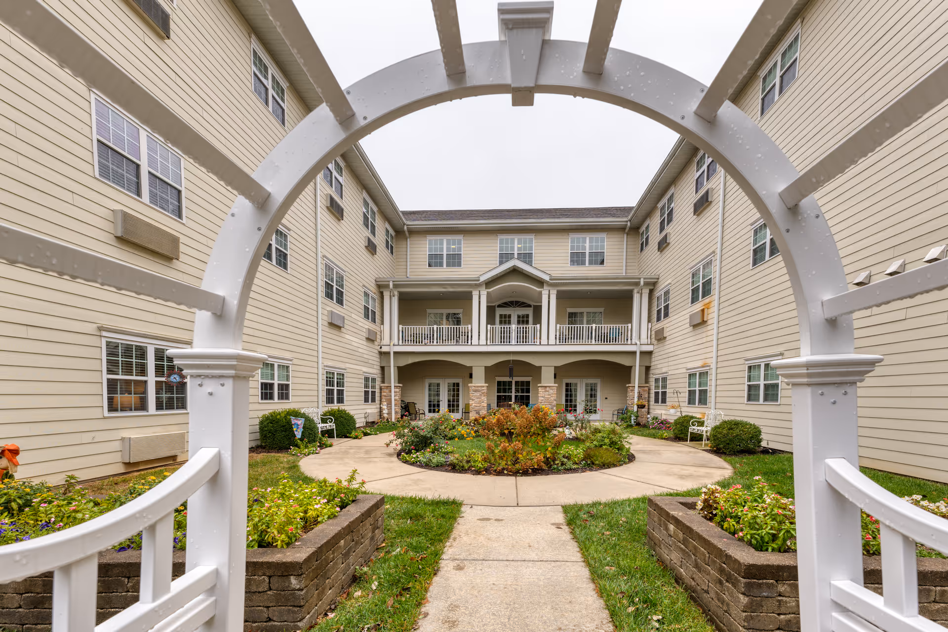 View through a white garden arch into a landscaped courtyard surrounded by a multi-story senior living building with balconies.