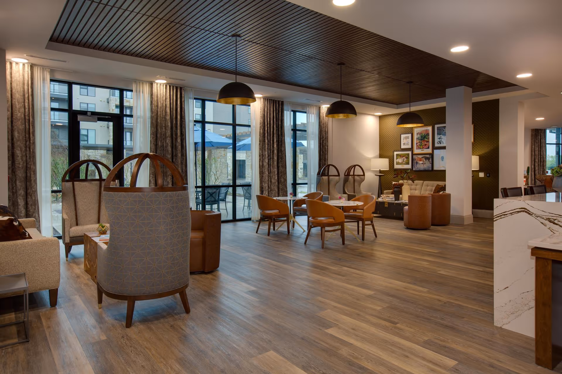 A spacious and modern living room area with wooden flooring, multiple seating arrangements including armchairs and small tables, large windows with curtains allowing natural light, pendant lights hanging from a dark wooden ceiling, and framed artwork on the walls.