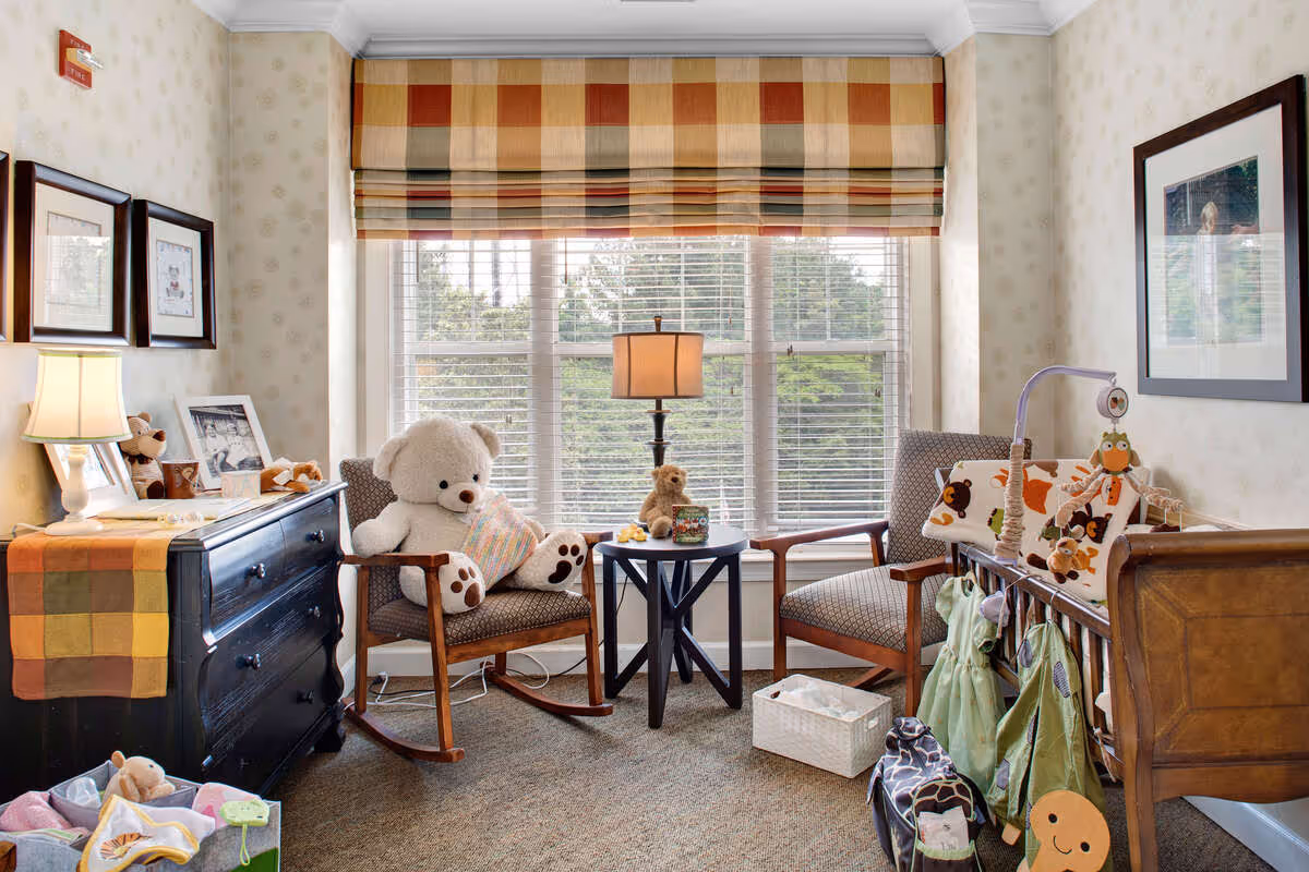 A cozy room with two cushioned wooden rocking chairs, one holding a large white teddy bear. Between the chairs is a small round table with a lamp and a small teddy bear. To the right is a wooden crib with colorful animal-themed bedding and hanging toys. On the left side, there is a black dresser with a lamp, framed pictures, and a colorful table runner. The room has a large window with striped blinds and light-patterned wallpaper.