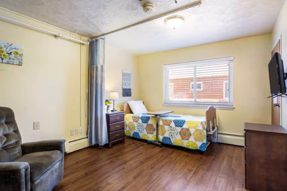 A small, cozy bedroom in a senior living facility with a single bed covered by a colorful quilt, a bedside table with a lamp and flowers, a recliner chair, a wall-mounted TV, and a window with blinds letting in natural light.