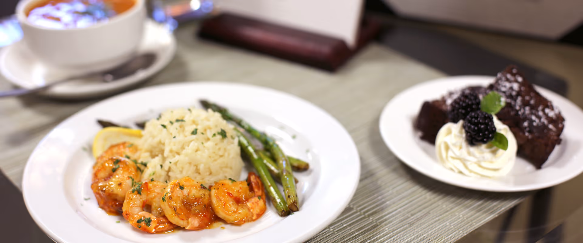 A plated meal featuring grilled shrimp, a serving of rice, and asparagus on a white plate, accompanied by a dessert plate with chocolate cake, whipped cream, blackberries, and a mint garnish, set on a table with a light-colored placemat.