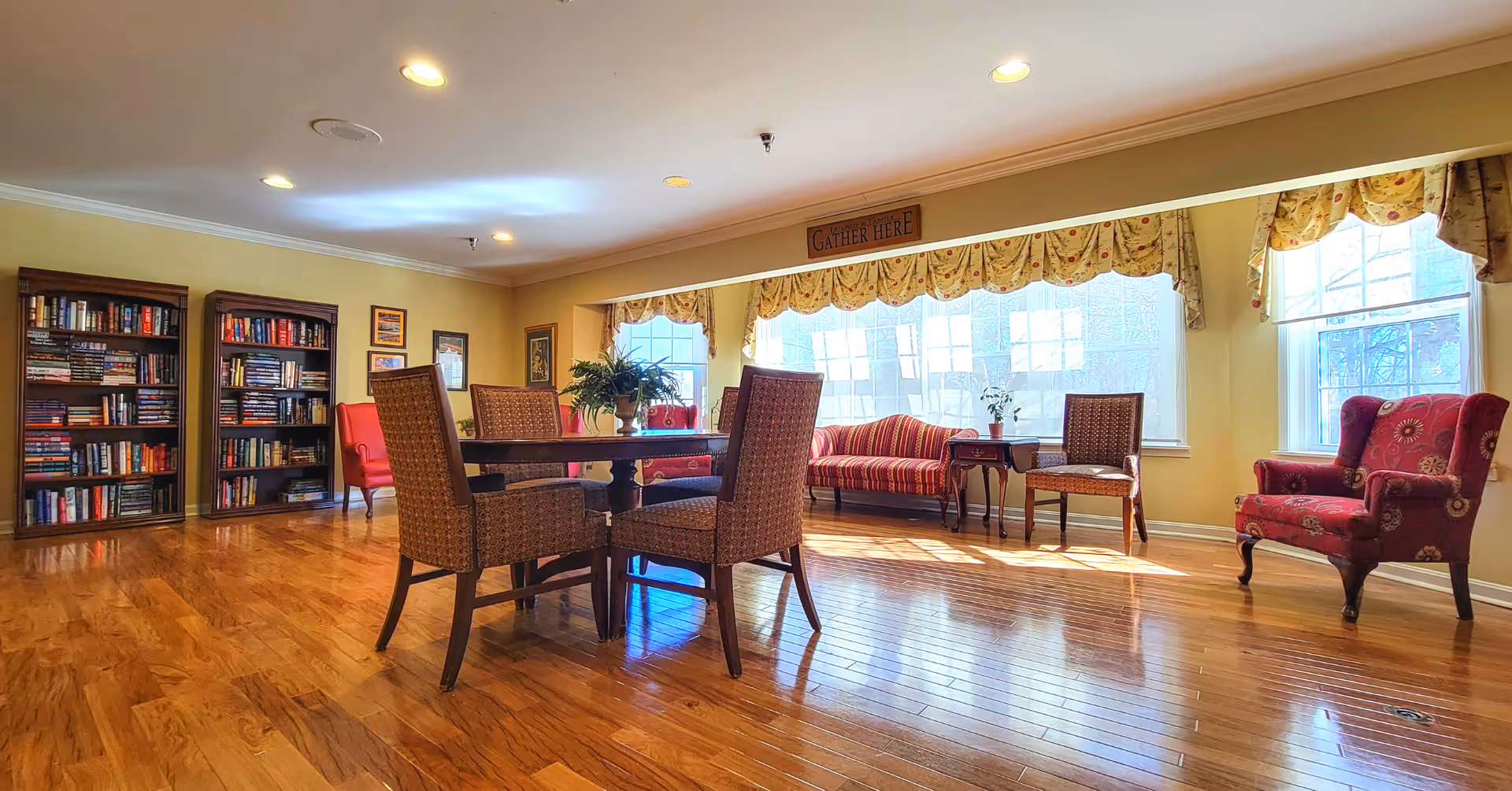 A bright and spacious common area with wooden flooring, featuring a round table with four cushioned chairs in the center. There are bookshelves filled with books on the left side, and several upholstered chairs and a small sofa near large windows with floral valances. A sign above the windows reads 'GATHER HERE'.