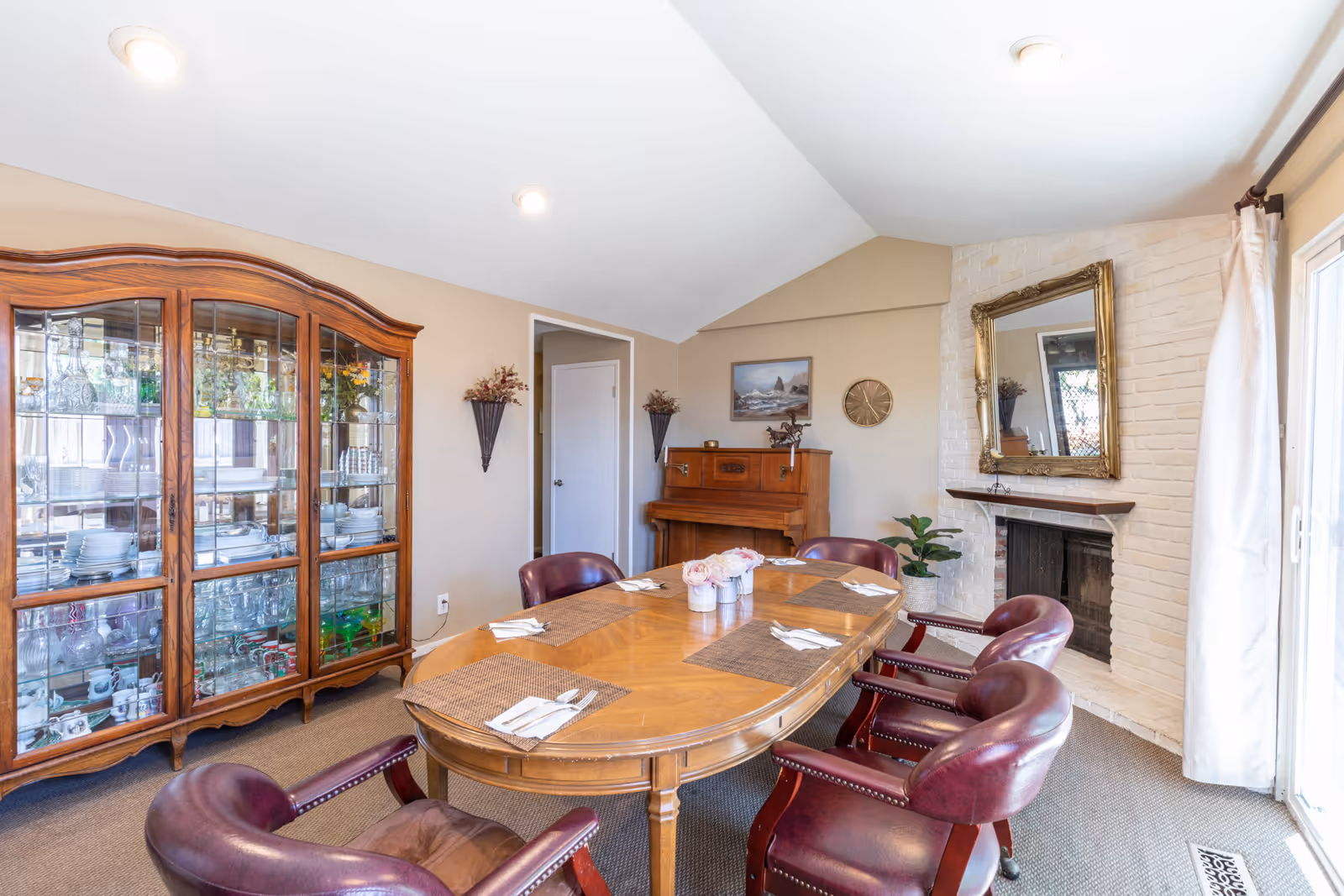 A bright dining room featuring a wooden oval table set with placemats and leather chairs, a glass-front china cabinet, a piano, and a fireplace with a mirror.