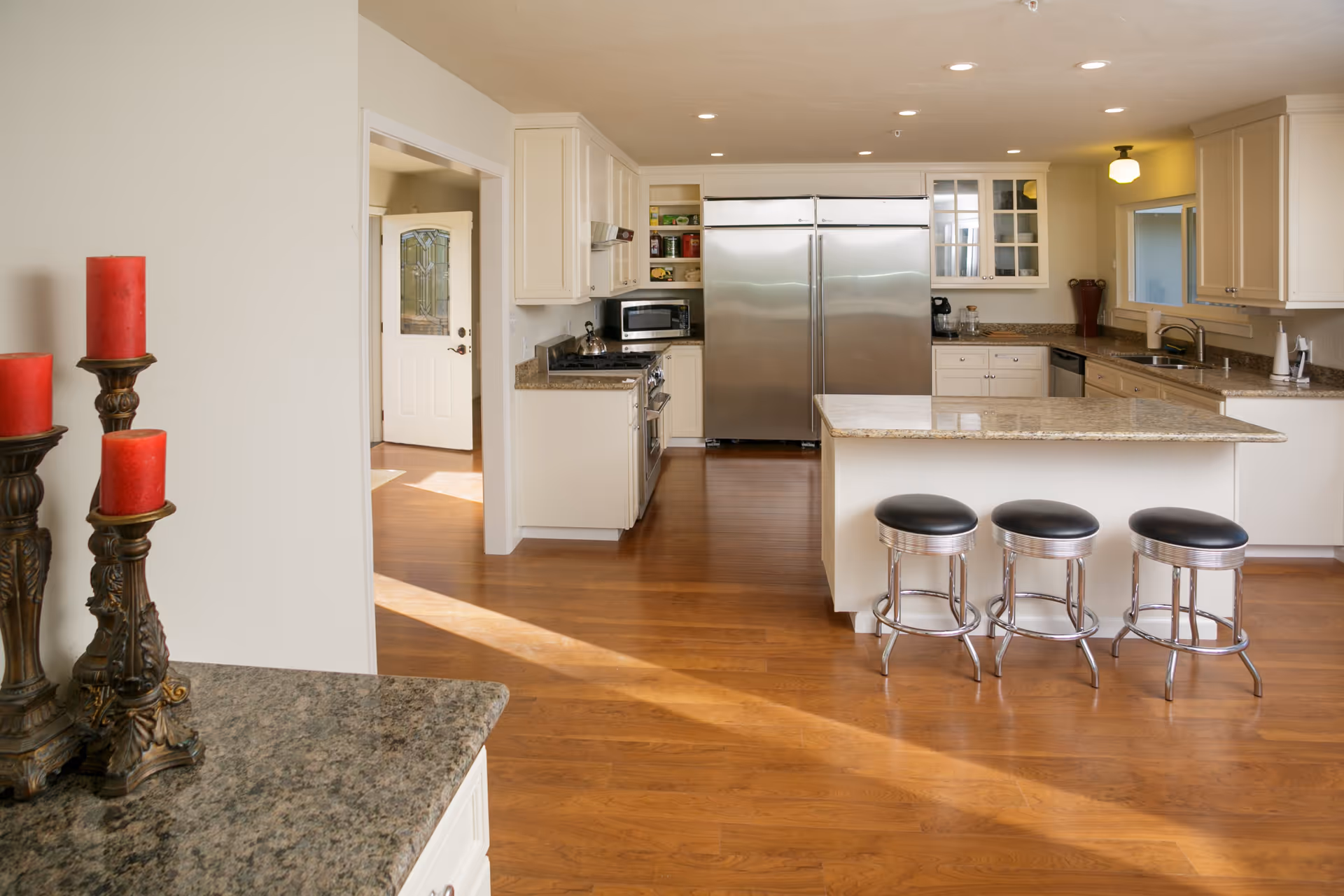 Bright open kitchen with a center island, three metal stools, stainless steel refrigerator and hardwood floors.